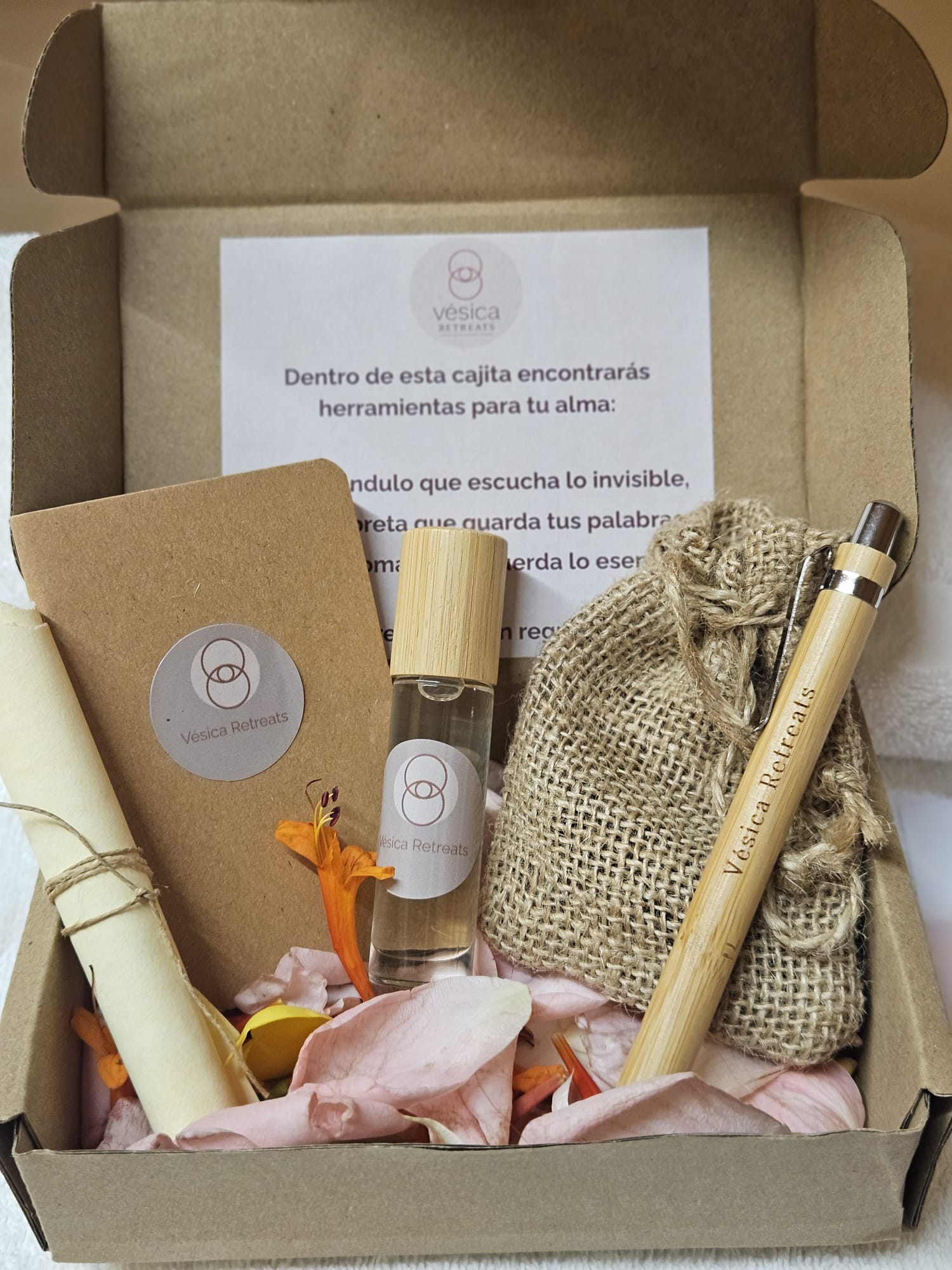 La cajita de bienvenida de Vésica abierta: una libreta, un péndulo, un frasco de aroma y un bolígrafo de bambú sobre pétalos