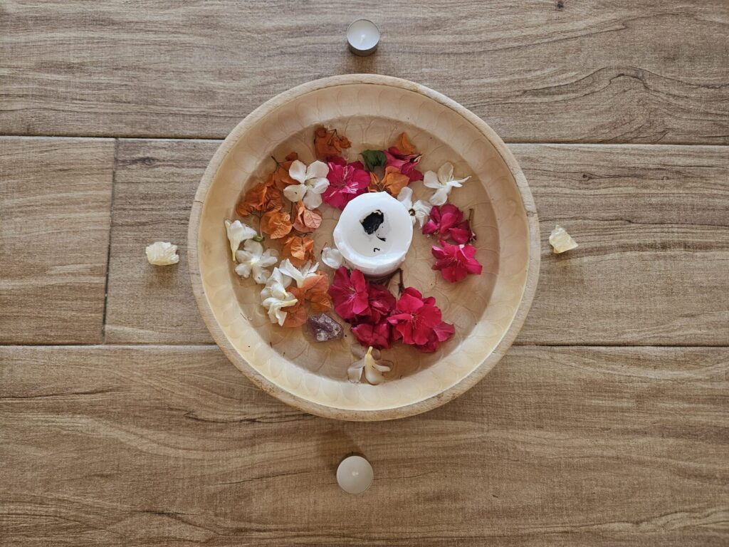 Un cuenco de madera con agua, pétalos de flores de colores y una vela blanca encendida en el centro, visto desde arriba sobre el suelo de madera