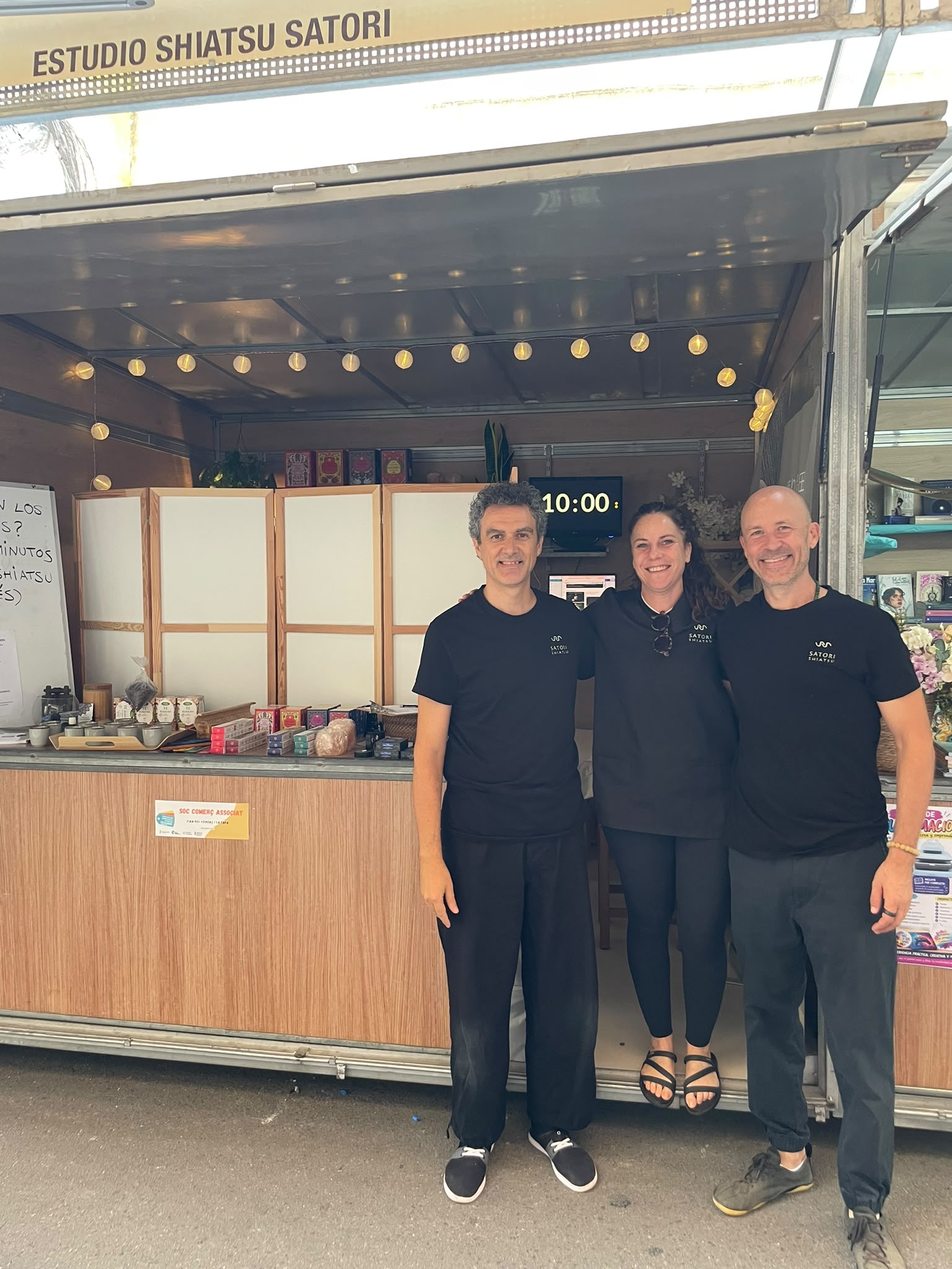 Tres personas del equipo de Estudio Shiatsu Satori, con camiseta negra, delante de la caseta de madera de la feria. Al fondo se ven los biombos y una pantalla con el cronómetro en diez minutos.