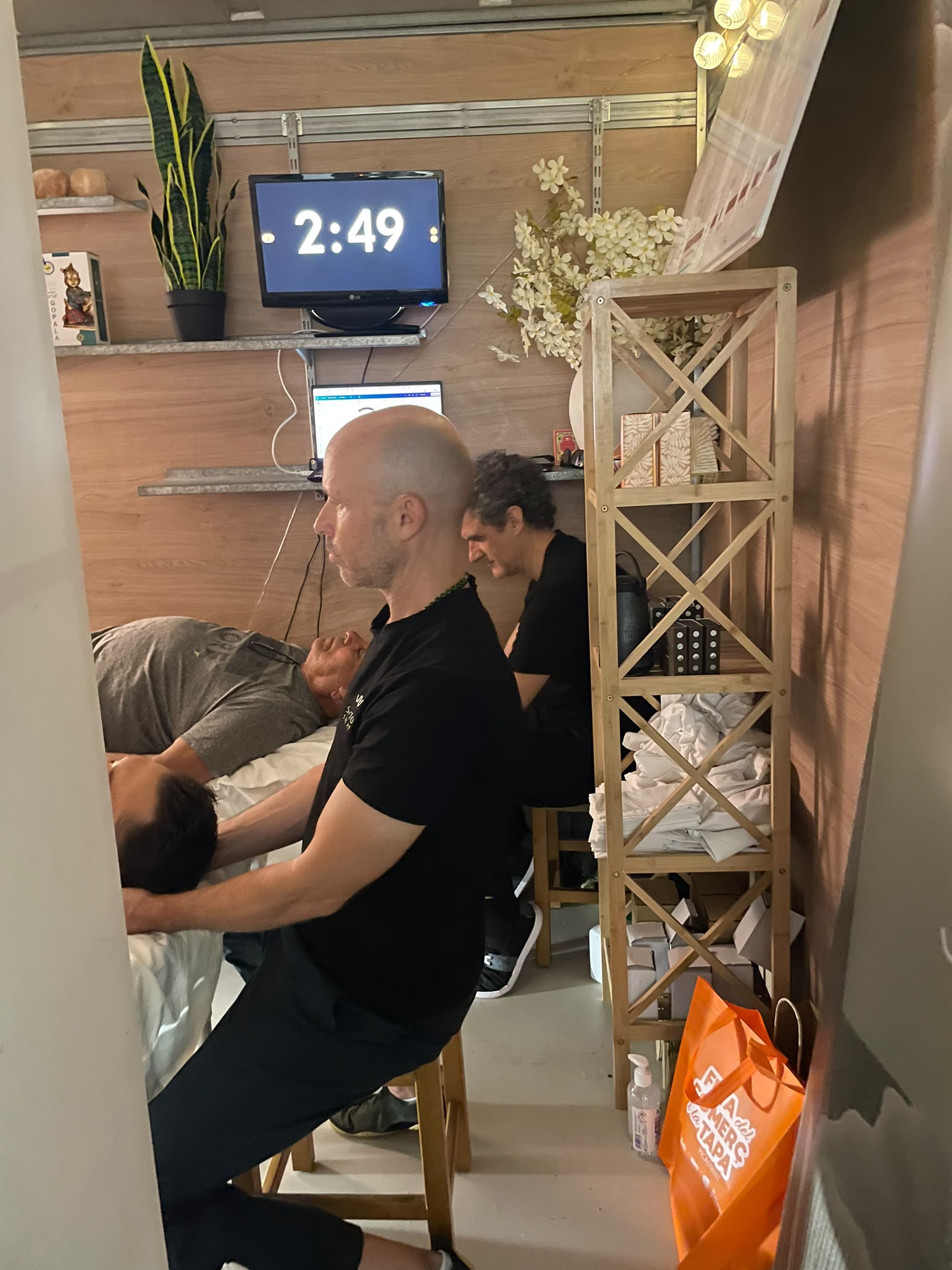 Dos terapeutas dando Shiatsu a dos personas tumbadas en camilla dentro de la caseta. En la pantalla del fondo, el cronómetro marca 2:49.